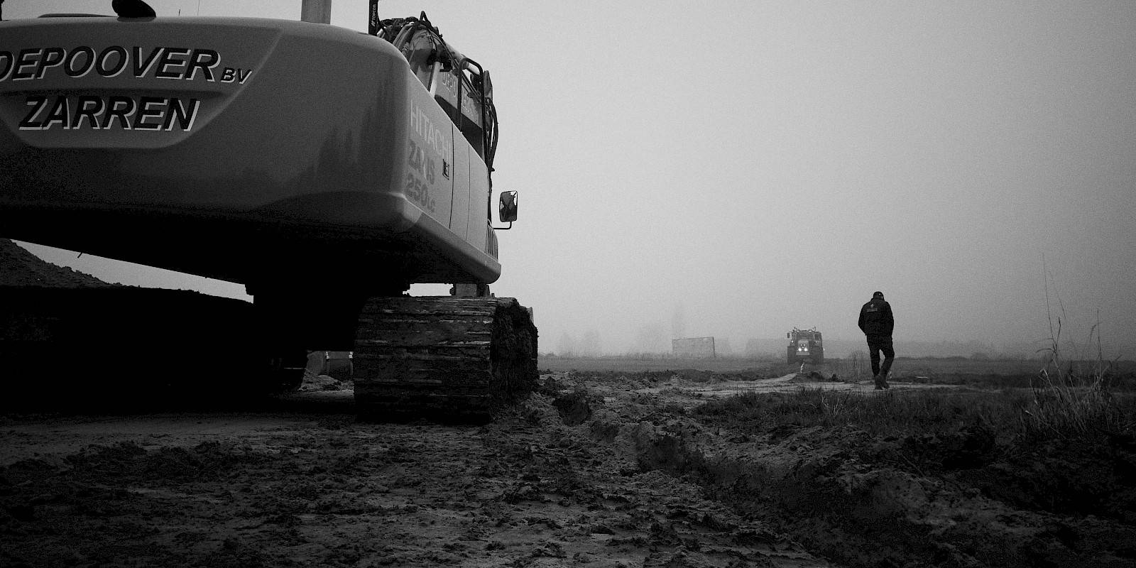 Photographie en noir et blanc d'une grue excavatrice dans un champ, avec un ouvrier et un tracteur à l'arrière-plan