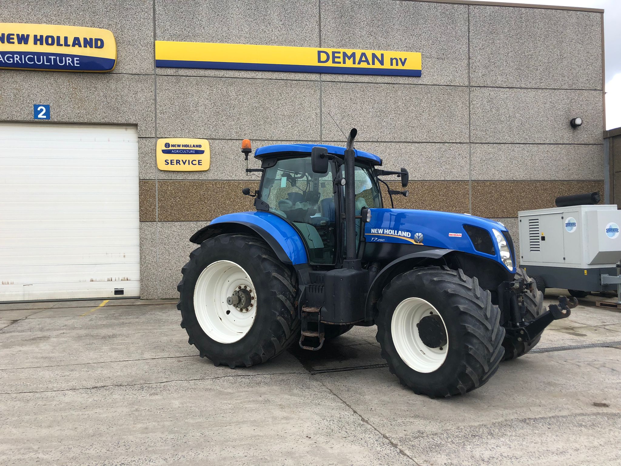 Een blauwe NEW HOLLAND T7.250 in zijaanzicht geparkeerd voor de de gebouwen van Deman nv